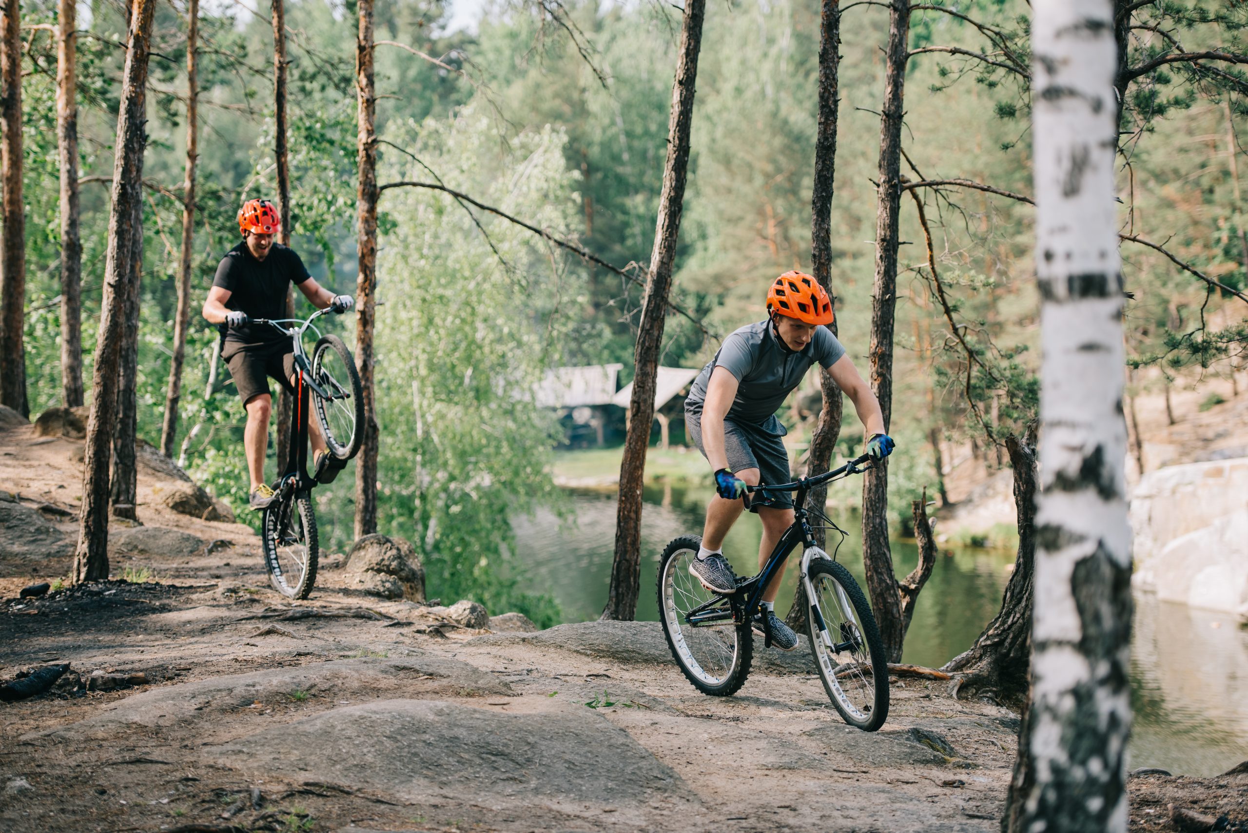male extreme cyclist in protective helmet balancing on back wheel of mountain bicycle while his friend riding near in forest